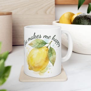 Könnte beinhalten: Weißer Keramikbecher mit einer Aquarellillustration einer Zitrone und Blättern. Der Becher trägt den Text "Tea makes me happy" in handschriftlichem Stil. Der Becher steht auf einem beigen Untersetzer.