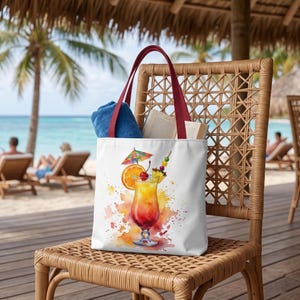 Könnte beinhalten: Eine weiße Tragetasche mit dunkelroten Henkeln, die eine Aquarellillustration eines tropischen Cocktails in einem Glas mit Orangenscheibe, Kirsche und kleinem bunten Schirmchen zeigt. Ein blaues Handtuch und ein offenes Buch stecken in der Tasche.