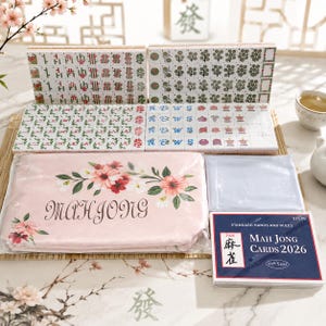 May include: A mahjong set featuring white tiles with green, red, and black symbols, a pink storage pouch with floral graphics and the word MAHJONG, and a blue instruction booklet titled Mah Jong Cards 2026 with standard hands and rules.