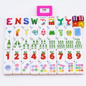 May include: A set of white mahjong tiles arranged in a grid, featuring colorful illustrations of letters, numbers, cocktails, beach items, and seashells. A pink tile holder with the letter E is at the top, alongside four translucent dice.