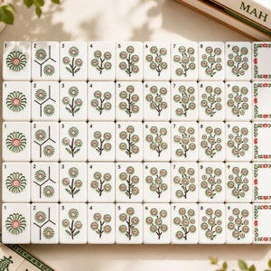 May include: A set of white Mahjong tiles arranged in a grid, featuring floral illustrations in green, pink, and black. Each tile displays a number from one to nine, with the final column showing blank tiles framed by green and red floral borders.