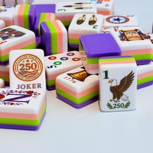May include: A collection of rectangular Mahjong tiles featuring a layered design with purple, lime green, and pink stripes. The tiles display various graphics including a United States 250th anniversary emblem, a bald eagle, a bell, and the text 1776-2026.