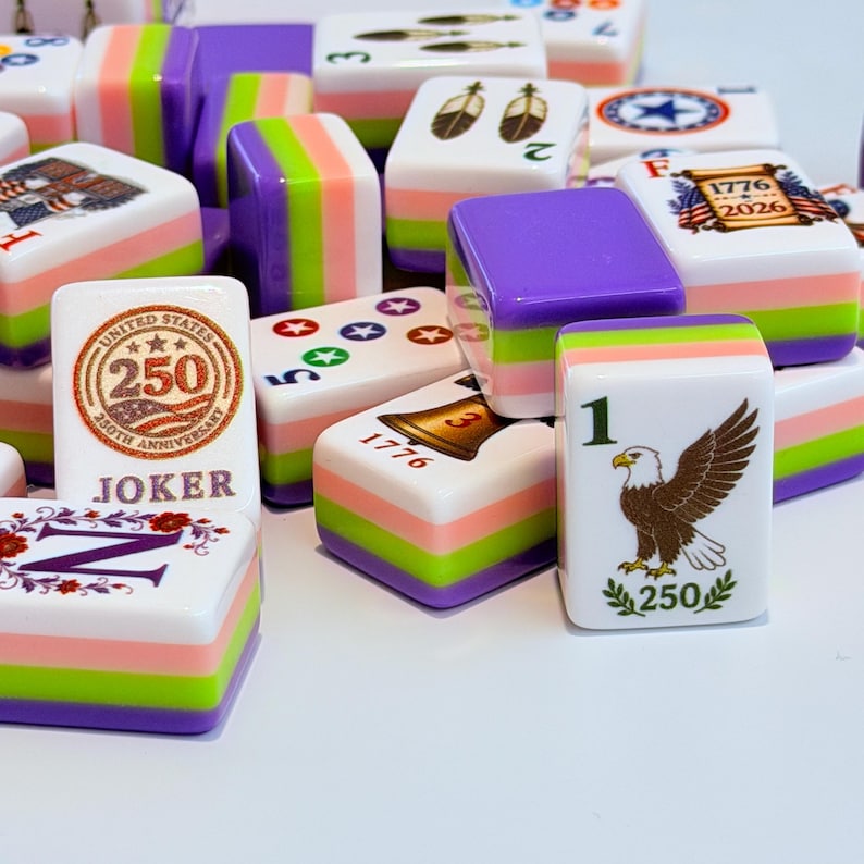 May include: A collection of rectangular Mahjong tiles featuring a layered design with purple, lime green, and pink stripes. The tiles display various graphics including a United States 250th anniversary emblem, a bald eagle, a bell, and the text 1776-2026.