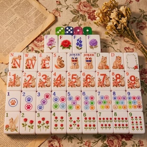 May include: A set of white mahjong tiles arranged in rows, featuring colorful illustrations of cats, flowers, paw prints, and numbers. The tiles include lettered markers, joker tiles, and numbered suits, all decorated with detailed floral and feline motifs.