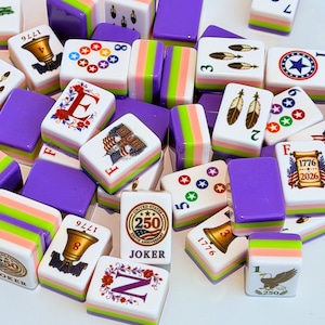 May include: A collection of rectangular mahjong tiles featuring white faces with colorful graphics including bells, feathers, stars, eagles, and text such as 1776, 2026, and 250th Anniversary Joker. The tiles have purple, green, and pink striped sides.