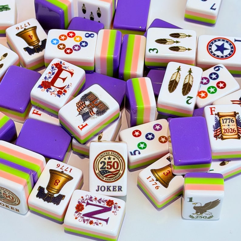 May include: A collection of rectangular mahjong tiles featuring white faces with colorful graphics including bells, feathers, stars, eagles, and text such as 1776, 2026, and 250th Anniversary Joker. The tiles have purple, green, and pink striped sides.