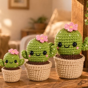 May include: Three crocheted cactus plants in beige pots. The cacti are green with small arms, black eyes, and pink flower accents. The pots are filled with brown material, and the background is blurred.