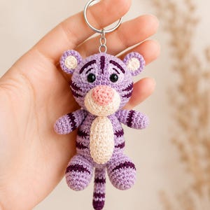 May include: A handmade crochet tiger keychain in shades of purple and cream. The tiger has dark purple stripes, a pink nose, and black eyes. The keychain is attached to a silver metal ring.