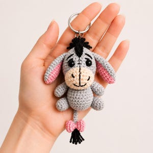 Peut inclure: Un porte-clés en crochet représentant un âne gris avec un museau beige clair, des cheveux en laine noire et l'intérieur des oreilles rose. L'âne a des yeux et une bouche brodés en noir, avec un petit nœud rose et un pompon noir sur la queue.