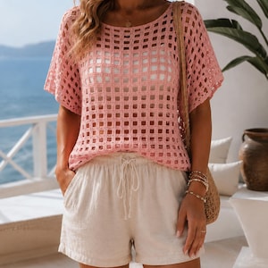 Può includere: Un top a maniche corte in crochet rosa con motivo a griglia quadrata e scollo rotondo, abbinato a pantaloncini beige con coulisse, vita elastica e tasche laterali. Il top presenta una lavorazione a maglia aperta e una vestibilità ampia.