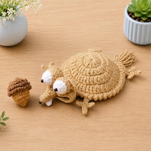 May include: A tan crocheted squirrel-like creature with large white eyes, black pupils, and a long snout, positioned next to a small brown crocheted acorn. The creature has a textured, rounded body, small limbs, and a short tail made of yarn.