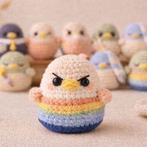 Pode incluir: Uma coleção de figuras de pássaros de crochê feitas à mão. O pássaro em primeiro plano tem uma expressão zangada, com cabeça branca, bochechas pêssego e corpo listrado em arco-íris. Outros pássaros no fundo têm várias cores e designs.