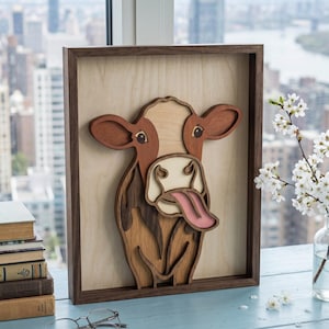 May include: A wooden cow art piece with a brown frame. The cow has a light brown and white face, brown ears, and a pink tongue sticking out. The artwork is displayed on a light wooden background.