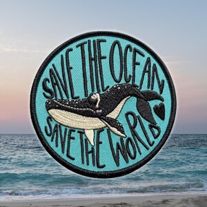May include: A circular embroidered patch with a light blue background featuring a black whale illustration in the center. The text "SAVE THE OCEAN" is arched above the whale, and "SAVE THE WORLD" is arched below, with a small black heart icon.