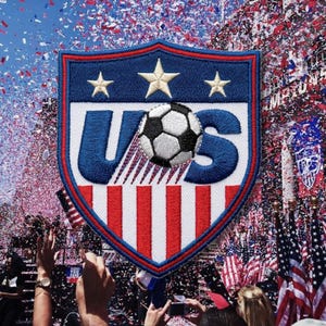 May include: A shield-shaped embroidered patch featuring the letters US in blue, a black and white soccer ball, three white stars, and red and white vertical stripes, set against a background of falling confetti and American flags.