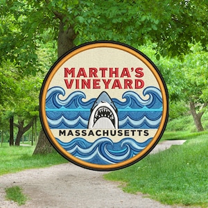 May include: Embroidered patch featuring a shark with an open mouth, set against blue waves. The text "MARTHA'S VINEYARD" arches above the shark, with "MASSACHUSETTS" below. The patch has a tan background and a black border.
