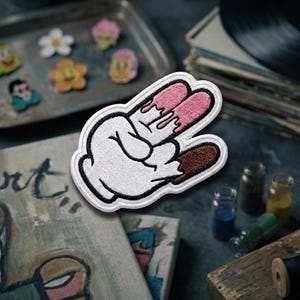May include: Embroidered patch featuring a white gloved hand making a peace sign with pink and brown dripping details. The patch has a black border. Other patches and art supplies are in the background.