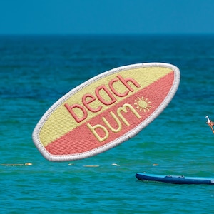 May include: Embroidered patch in the shape of a surfboard, with a yellow and red color scheme. The text "beach bum" is embroidered in pink, with a small sun graphic. The patch is floating above the water.