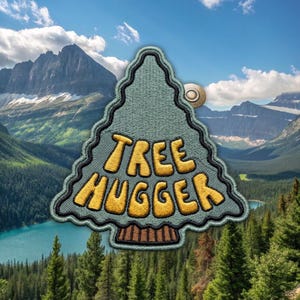 May include: A triangular embroidered patch shaped like a pine tree with a sage green background, a brown trunk, and a black border. The text "TREE HUGGER" is stitched in bold, yellow, rounded letters across the center of the patch.