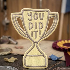 May include: A yellow and gray embroidered patch in the shape of a trophy. The patch features the words "YOU DID IT!" in gray lettering. The trophy has a classic design with a base and handles. The background includes medals and ribbons.
