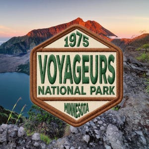 May include: Embroidered patch featuring the text "VOYAGEURS NATIONAL PARK MINNESOTA" in green and white, with the year "1975" above. The patch has a brown border and is set against a scenic mountain and lake backdrop.