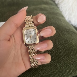 Può includere: Un orologio da polso color oro con quadrante rettangolare e cinturino a maglie. Il quadrante dell'orologio è argentato con lancette nere e il marchio "XIMILI" è visibile. L'orologio è tenuto in una mano con unghie lunghe e lucide.