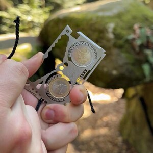 May include: A silver multi-tool with two coins, held against a blurred outdoor background. The tool has various cutouts and a ruler edge. A black cord is held in the other hand.