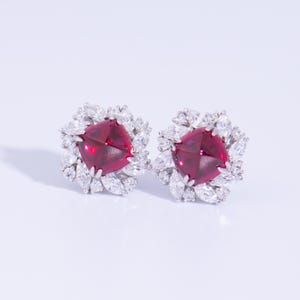 May include: A pair of elegant stud earrings. Each earring showcases a central, square, deep red gemstone, encircled by smaller, clear, marquise-cut stones. The earrings are set against a white backdrop, accentuating their brilliance.