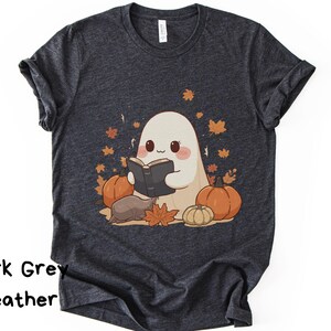 Puede incluir: Una camiseta de manga corta en color gris jaspeado oscuro con un gráfico de un fantasma blanco leyendo un libro, rodeado de calabazas naranjas y hojas de otoño dispersas. El diseño incluye una pequeña criatura marrón junto al fantasma.