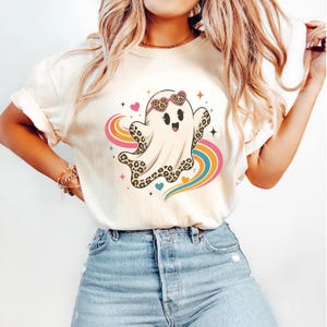 Puede incluir: Una camiseta de color crema con un gráfico de un fantasma blanco con detalles de estampado de leopardo, que lleva gafas de sol en forma de corazón. El fantasma está rodeado por un arcoíris colorido, corazones rosas y azules y estrellas.