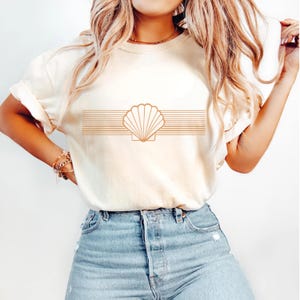 Puede incluir: Camiseta color crema con un diseño de concha. El diseño es una concha de color marrón claro con líneas horizontales arriba y abajo. La camiseta tiene mangas enrolladas y se combina con unos vaqueros azules.