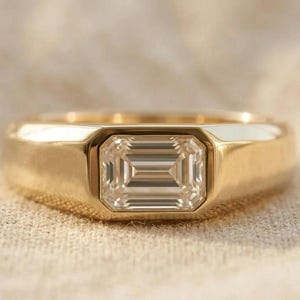 Könnte beinhalten: Ein goldener Ring mit einem rechteckigen Diamanten im Smaragdschliff. Der Ring hat ein dickes Band und eine Lünettenfassung. Der Diamant steht vor einem neutralen Hintergrund im Mittelpunkt.