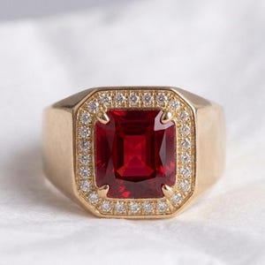 Könnte beinhalten: Ein Goldring mit einem großen, rechteckigen, tiefroten Edelstein in der Mitte. Der zentrale Stein ist von einem Kranz aus kleinen, klaren, rund geschliffenen weißen Steinen umgeben. Die Ringschiene ist aus glattem, poliertem Gold gefertigt.