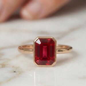 Könnte beinhalten: Ein Goldring mit einem großen, rechteckigen, im Smaragdschliff geschliffenen roten Edelstein, der in einer glatten, polierten Zargenfassung sitzt. Die Ringschiene ist schmal und aus passendem Goldmetall gefertigt, was den tiefroten Stein betont.