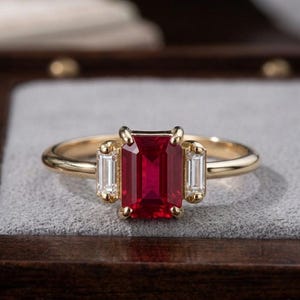 Könnte beinhalten: Ein Goldring mit einem zentralen, smaragdgeschliffenen roten Edelstein, der von zwei kleineren, im Baguetteschliff geschliffenen, klaren weißen Steinen flankiert wird. Der Ring hat eine polierte Goldschiene und eine Vier-Krappen-Fassung.