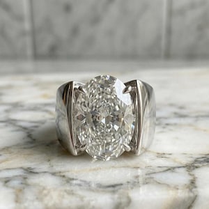 May include: A large, oval-cut diamond ring with a bright, clear appearance. The diamond is set in a wide, polished silver band. The ring is displayed on a marble surface.