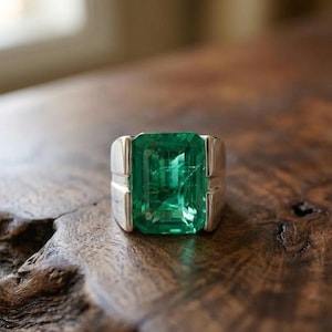 Könnte beinhalten: Ein Silberring mit einem großen, rechteckigen Smaragd-Edelstein. Der Smaragd hat eine leuchtend grüne Farbe und ist in einem schlichten Silberband gefasst. Der Ring wird auf einer Holzoberfläche präsentiert.