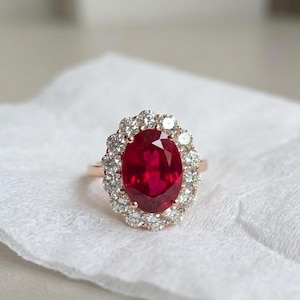 Könnte beinhalten: Ein roségoldener Ring mit einem großen, oval geschliffenen Rubin, umgeben von einem Kranz aus kleineren, klaren Edelsteinen. Der Rubin hat eine tiefrote Farbe, und die kleineren Steine funkeln. Der Ring liegt auf einer weißen Oberfläche.