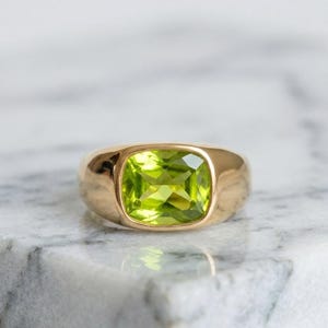 Könnte beinhalten: Ein goldener Ring mit einem großen, quadratisch geschliffenen Peridot-Edelstein. Der Peridot hat eine leuchtend hellgrüne Farbe und ist in einem glatten, polierten Goldband gefasst. Der Ring hat ein klassisches, elegantes Design.