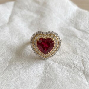 Könnte beinhalten: Ein herzförmiger Ring mit einem großen, tiefroten Edelstein in der Mitte. Der Edelstein ist von einem Kranz aus kleineren, klaren Steinen umgeben, die in einem goldenen Band gefasst sind. Der Ring liegt auf einer weißen, strukturierten Oberfläche.
