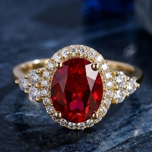 Op de afbeelding: Een gouden ring met een grote, ovaal geslepen, dieprode edelsteen, omringd door een halo van kleine, heldere diamanten. Aan weerszijden van de centrale steen zijn extra diamanten gezet, wat een sprankelend, elegant ontwerp creëert.