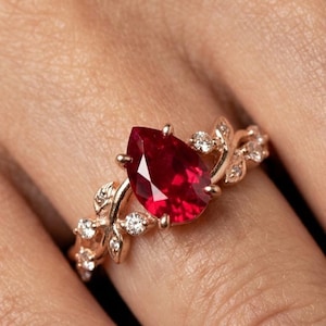 Könnte beinhalten: Ein roségoldener Ring mit einem birnenförmigen, tiefroten Edelstein. Der Ring ist mit kleinen, klaren Akzentsteinen und einem zarten, blattartigen Design verziert. Der Ring wird an einem Finger gezeigt.
