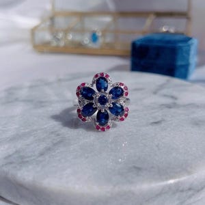 Könnte beinhalten: Ein silberner Ring mit Blumenmuster. Der Ring hat eine zentrale Anordnung kleiner, runder blauer Edelsteine, umgeben von größeren, ovalen blauen Edelsteinen. Die Blütenblätter sind mit kleinen, runden roten Edelsteinen eingefasst. Der Ring liegt auf einer weißen Marmoroberfläche.