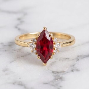 Könnte beinhalten: Ein Goldring mit einem zentralen, tiefroten Edelstein im Marquise-Schliff, der von Krappen gehalten wird. Der Mittelstein ist auf beiden Seiten von einer Gruppe aus drei kleinen, runden, klaren weißen Diamanten in Goldfassung umgeben.