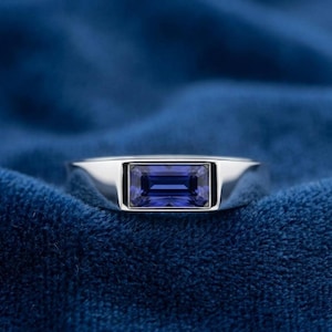 Könnte beinhalten: Ein silberner Ring mit einem rechteckigen, tiefblauen Edelstein. Der Ring hat ein schlichtes, modernes Design mit poliertem Finish. Der Edelstein ist in einer Lünettenfassung gefasst, auf blauem Samt.
