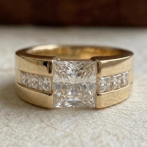 Könnte beinhalten: Ein goldener Ring mit einem großen, quadratisch geschliffenen Diamanten als Mittelstein. Kleinere quadratische Diamanten sind auf beiden Seiten des Mittelsteins gesetzt. Der Ring hat ein breites Band und eine polierte Oberfläche, die das Funkeln der Edelsteine hervorhebt.
