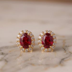 Könnte beinhalten: Ein Paar ovale Ohrstecker mit einem zentralen, tiefroten Rubin-Edelstein, der von einem Heiligenschein aus kleinen, klaren, rund geschliffenen weißen Diamanten umgeben ist, alles gefasst in einen Rahmen aus poliertem Gelbgold.