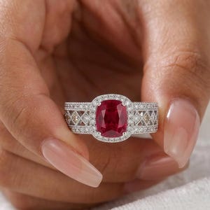 May include: A silver-toned ring featuring a large, cushion-cut, deep red gemstone at the centre, surrounded by a halo of small white stones. The band has a wide, intricate lattice design encrusted with numerous tiny, sparkling white stones.