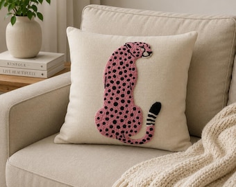 May include: A square throw pillow with a cream-colored fabric cover featuring a textured, embroidered pink cheetah with black spots. The cheetah is depicted in a seated profile position with a long tail that has black and white horizontal stripes.