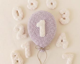 Puede incluir: Un globo de punch needle color lavanda con un número uno blanco en el centro, rodeado de números individuales blancos del cero al nueve dispuestos en un patrón circular, con un cordel fino de color marrón atado.